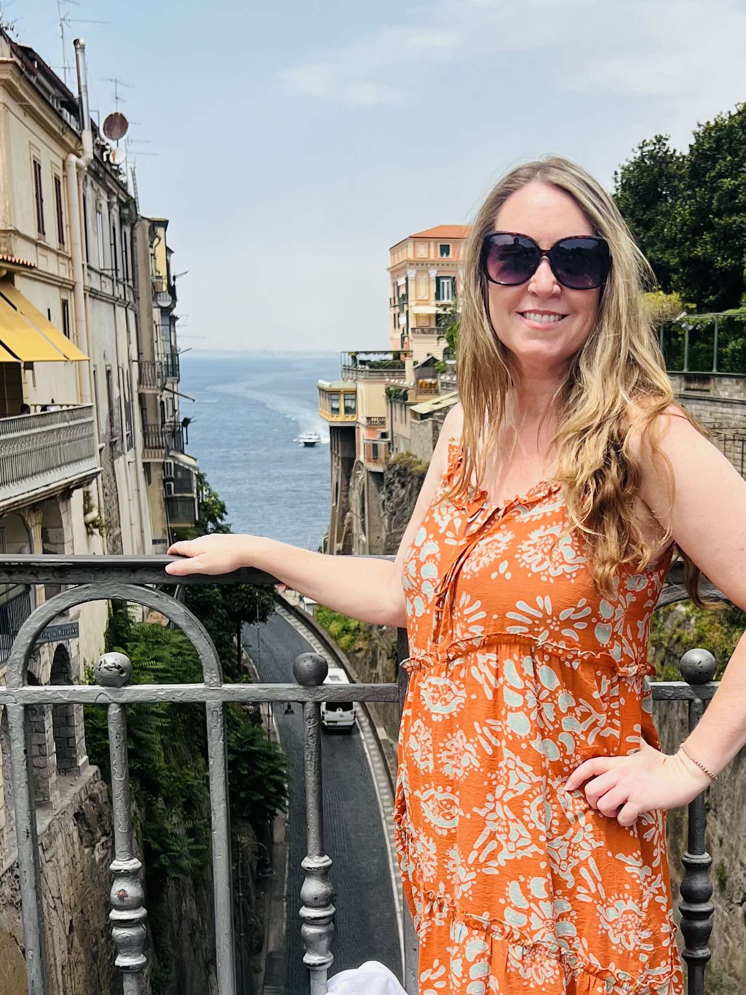 Me, a woman with long blonde hair in sundress and sunglasses in Sorrento Italy, posing with the cliffs and Bay of Naples in the background.