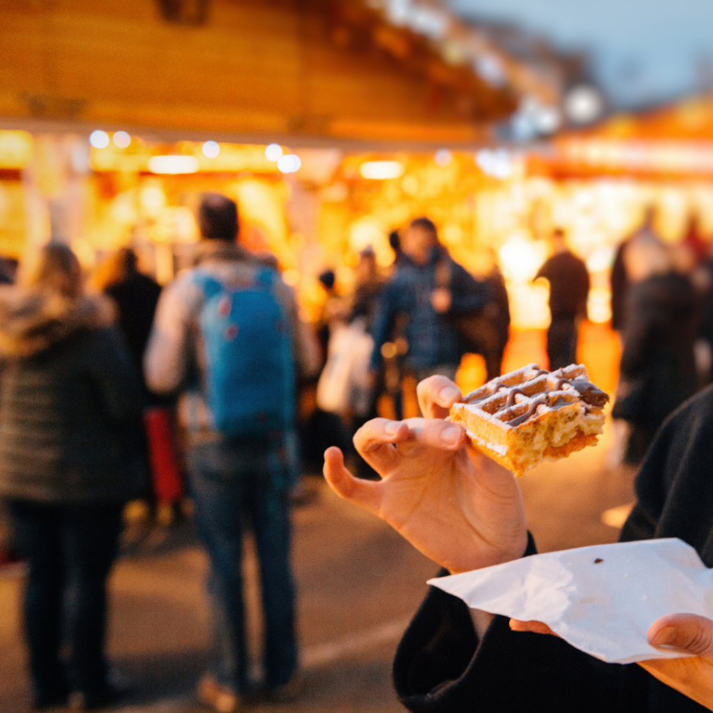 A person holding a waffle with nuetella drizzled on top with the christmas market buildings in the background.