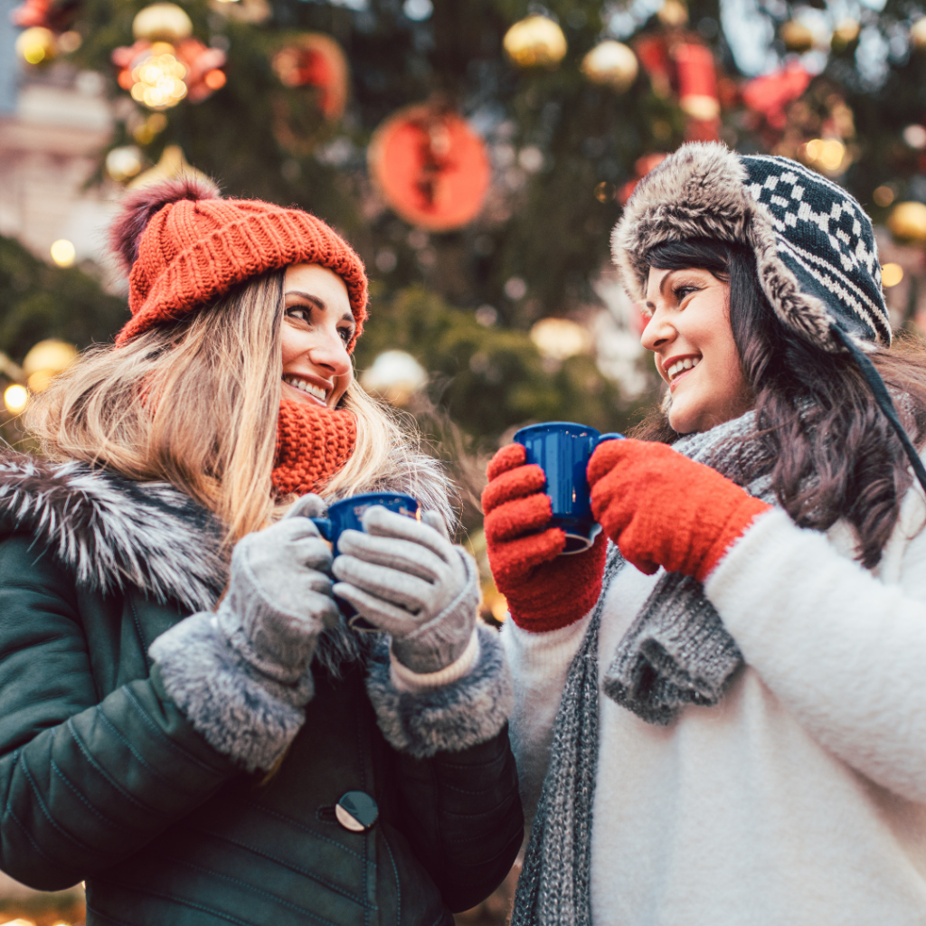 Two women dressed in winter coats and hats drinking from mugs and smiling in front of a Christmas tree