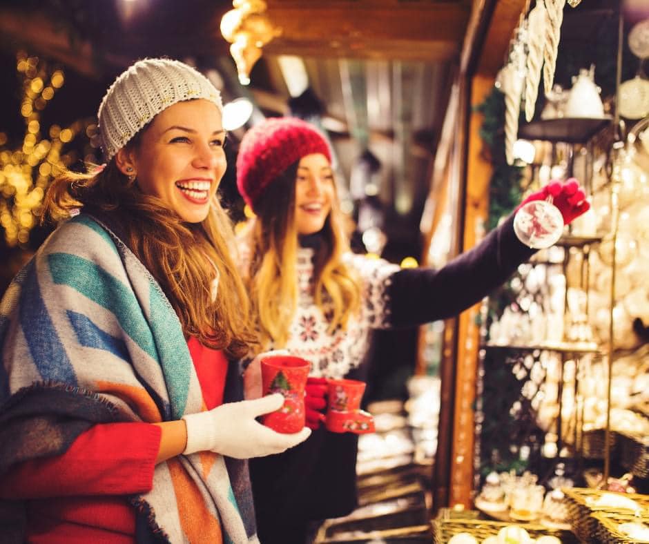 Two women smiling and holding a warm cup of mulled wine looking at decorations at a christmas markets stall.